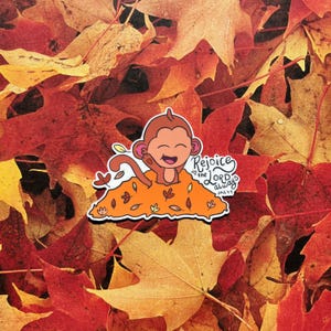 May include: A sticker featuring a cartoon monkey emerging from a pile of orange leaves, with the text "Rejoice in the Lord always phil 4:4". The sticker is set against a background of colorful autumn leaves.