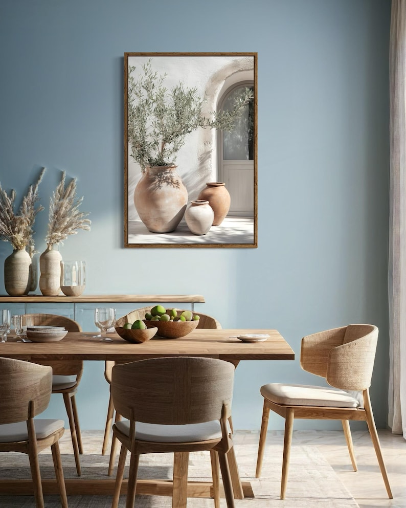 May include: A framed print of a whitewashed wall with an arched doorway and three terracotta pots with green plants. The pots are arranged in a row, with the largest pot in the center and the two smaller pots on either side. The print is hung on a light blue wall in a dining room with a wooden table and chairs.