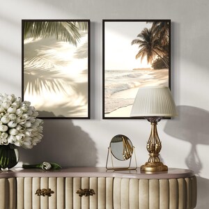 Puede incluir: Dos impresiones enmarcadas de escenas de playa con palmeras y olas del oc&eacute;ano, una l&aacute;mpara de mesa dorada con una pantalla blanca, un peque&ntilde;o espejo redondo y un jarr&oacute;n de flores blancas sobre una mesa beige con detalles dorados.