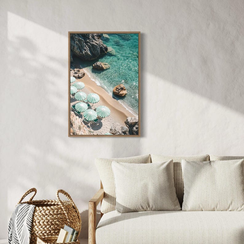 May include: A framed print of a beach scene with turquoise water, a sandy shore, and green and white striped umbrellas. Large rocks are visible in the water and on the beach. The print is displayed on a white wall.
