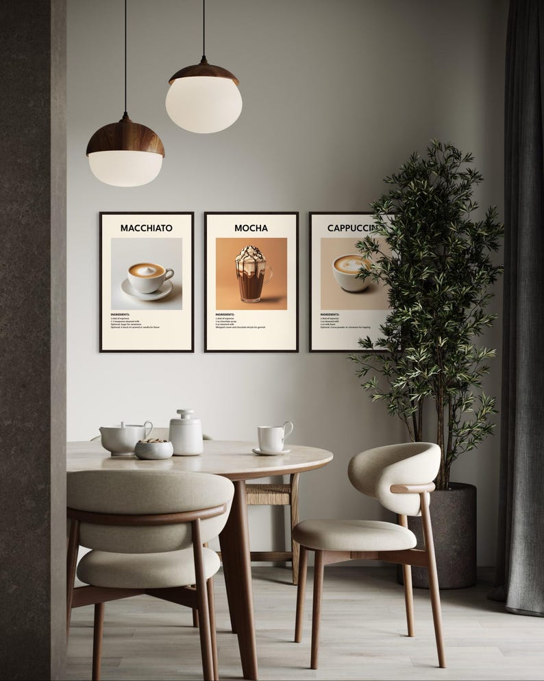 May include: Three framed prints of coffee drinks with the names "Macchiato", "Mocha", and "Cappuccino" printed below each image. The prints are hanging on a light gray wall above a wooden table with two chairs.