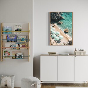 May include: A white cabinet with three doors and gold handles. The cabinet is sitting on a wooden floor. There is a white sheep-shaped ottoman in front of the cabinet. A rug with a black and white pattern is on the floor. A framed print of a beach scene with turquoise water and white umbrellas is hanging on the wall above the cabinet.