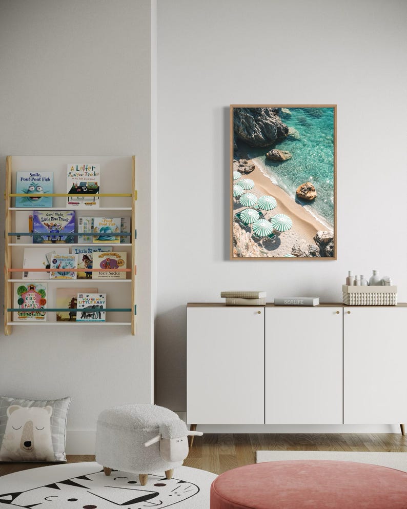 May include: A white cabinet with three doors and gold handles. The cabinet is sitting on a wooden floor. There is a white sheep-shaped ottoman in front of the cabinet. A rug with a black and white pattern is on the floor. A framed print of a beach scene with turquoise water and white umbrellas is hanging on the wall above the cabinet.
