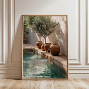 May include: A framed print featuring a tranquil outdoor scene. A rectangular pool of turquoise water is in the foreground. Brown terracotta pots with water spouts line the edge of the pool. An olive tree and a white wall are in the background.