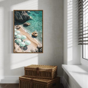 May include: A framed print of a beach scene with turquoise water, white sand, and green and white striped umbrellas. Two woven storage trunks are stacked below the framed print. The scene is illuminated by natural light from a nearby window.