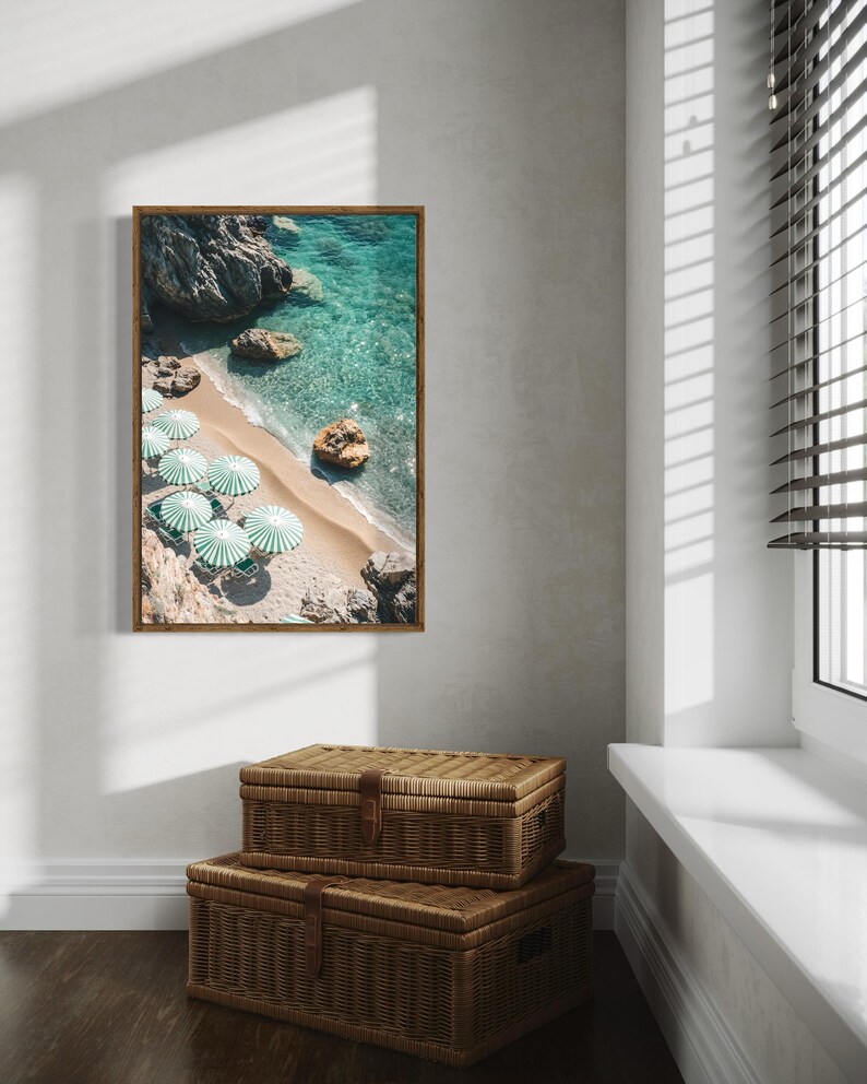 May include: A framed print of a beach scene with turquoise water, white sand, and green and white striped umbrellas. Two woven storage trunks are stacked below the framed print. The scene is illuminated by natural light from a nearby window.