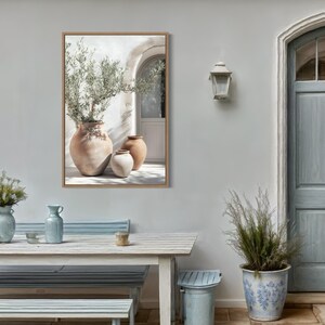 May include: A framed print of three terracotta pots with a green plant in the largest pot. The pots are sitting on a white tiled floor in front of a white wall with a doorway. The print is hanging on a light gray wall.