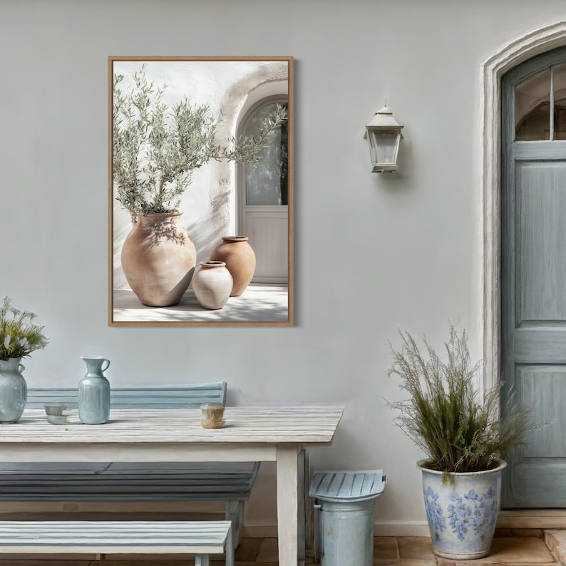 May include: A framed print of three terracotta pots with a green plant in the largest pot. The pots are sitting on a white tiled floor in front of a white wall with a doorway. The print is hanging on a light gray wall.
