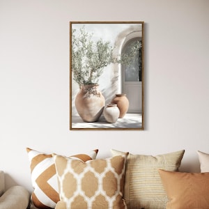 May include: A framed print of a white building with a doorway and three terracotta pots with olive branches. The pots are arranged in a row on the ground in front of the building. The print is set against a white wall with a woven light fixture hanging above.