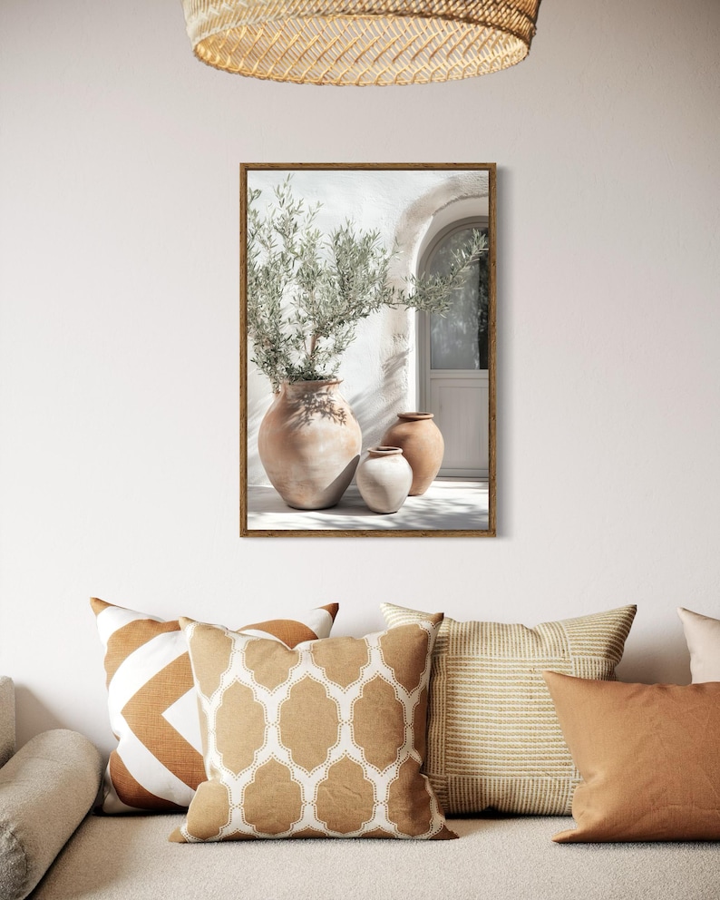 May include: A framed print of a white building with a doorway and three terracotta pots with olive branches. The pots are arranged in a row on the ground in front of the building. The print is set against a white wall with a woven light fixture hanging above.