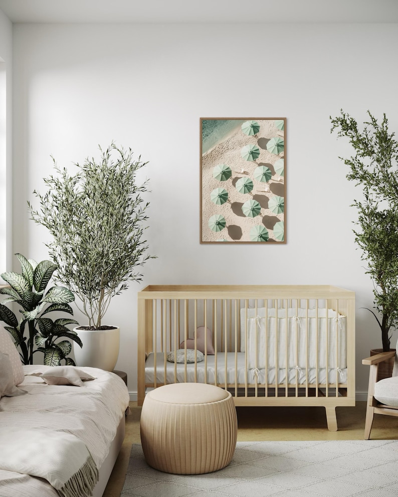 May include: A framed print of an aerial view of a beach with green umbrellas and white sand. The print is hanging on a white wall in a nursery with a crib and a plant.