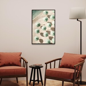 May include: A framed print of a beach scene with green umbrellas arranged in rows on the sand. The print is hanging on a white wall above two orange armchairs and a black side table.