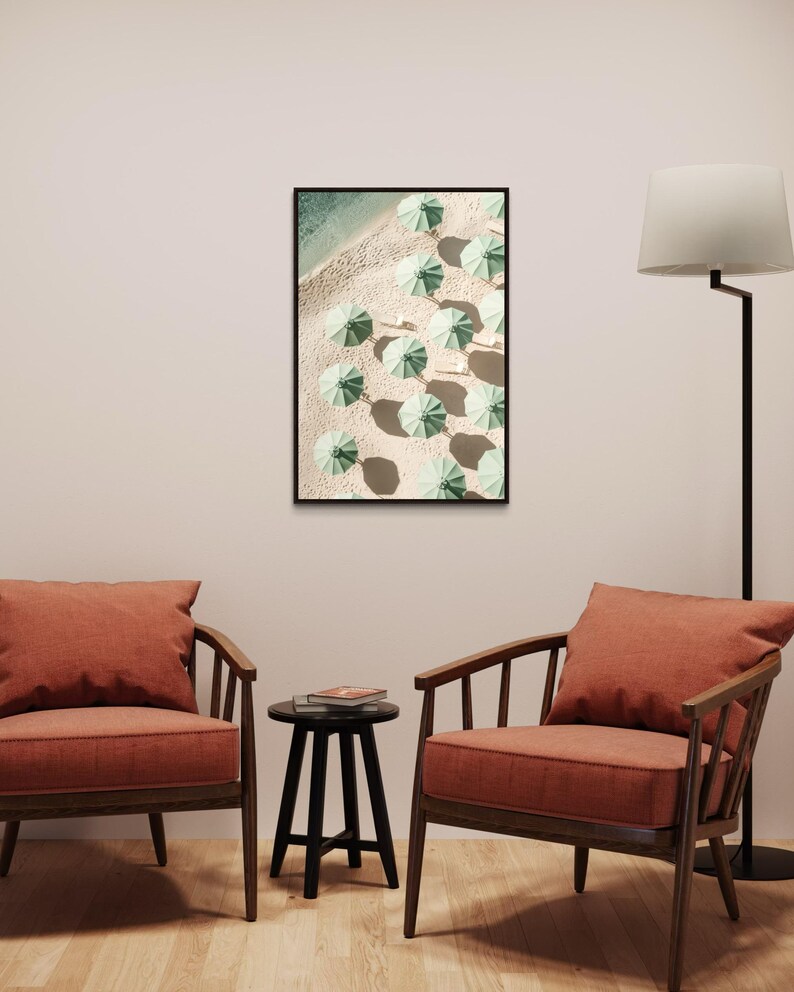 May include: A framed print of a beach scene with green umbrellas arranged in rows on the sand. The print is hanging on a white wall above two orange armchairs and a black side table.
