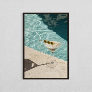 Könnte beinhalten: Ein Martini-Glas gefüllt mit einer klaren Flüssigkeit und mit drei grünen Oliven an einem Zahnstocher garniert. Das Glas steht auf einer hellbeigen Oberfläche neben einem blauen Schwimmbecken.