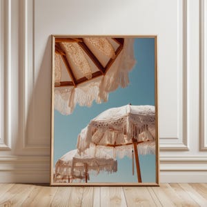 May include: A framed print featuring three white, fringed parasols against a clear blue sky. The parasols have wooden frames and intricate lace detailing. The image evokes a sense of relaxation and leisure.