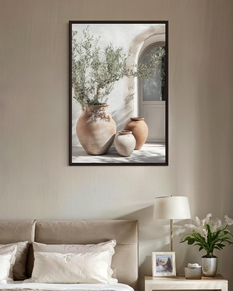 May include: A framed print of a white wall with an arched doorway and three terracotta pots. The pots are arranged in a row, with a small olive tree in the largest pot. The print has a rustic and minimalist aesthetic.