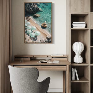 May include: A framed print of a beach scene with turquoise water, a sandy shore, and several green and white striped umbrellas. The print hangs above a wooden desk with a gray chair.