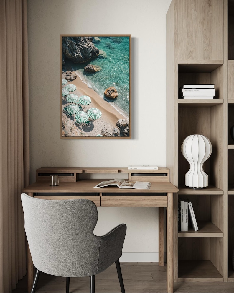 May include: A framed print of a beach scene with turquoise water, a sandy shore, and several green and white striped umbrellas. The print hangs above a wooden desk with a gray chair.