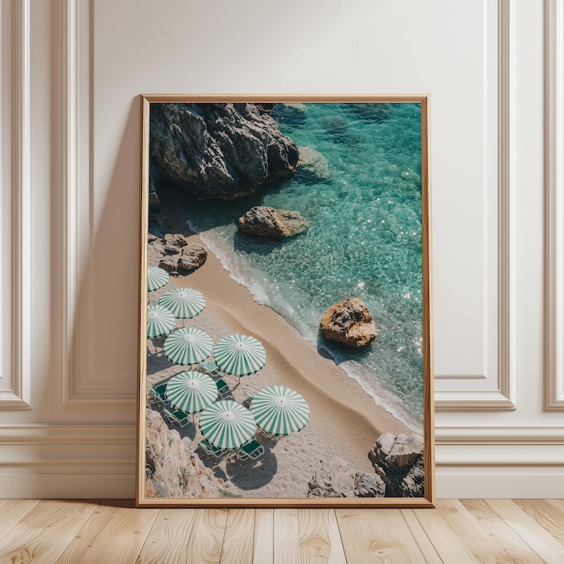 May include: A framed print featuring a beach scene with turquoise water, sandy shore, and striped green and white umbrellas. The image includes rocks, beach chairs, and a view of the ocean. The print is displayed against a white wall and wooden floor.