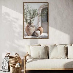 May include: A framed print of a white wall with a terracotta pot containing a green plant and two smaller terracotta pots. The print is hanging on a white wall above a light brown couch with white cushions.