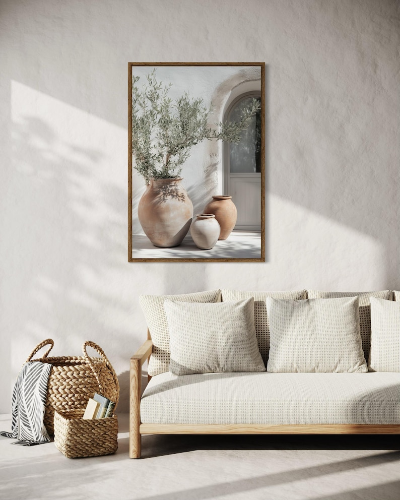 May include: A framed print of a white wall with a terracotta pot containing a green plant and two smaller terracotta pots. The print is hanging on a white wall above a light brown couch with white cushions.