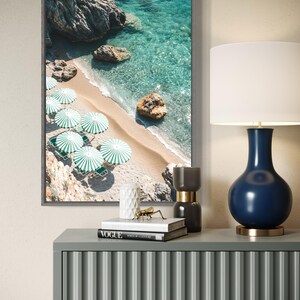May include: A framed photograph of a beach scene with turquoise water, white sand, and green umbrellas. The photo is displayed on a gray console table with a blue and gold lamp, books, and decorative objects.