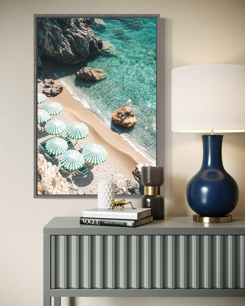 May include: A framed photograph of a beach scene with turquoise water, white sand, and green umbrellas. The photo is displayed on a gray console table with a blue and gold lamp, books, and decorative objects.
