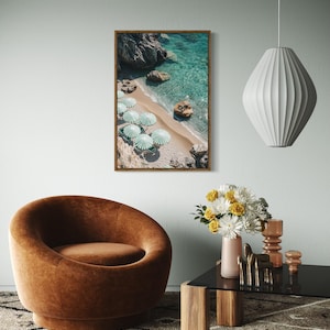 May include: A framed print of a beach scene with turquoise water, sandy shore, and green and white striped umbrellas. A brown velvet armchair, a black glass table, and a white pendant lamp are also visible in the room.