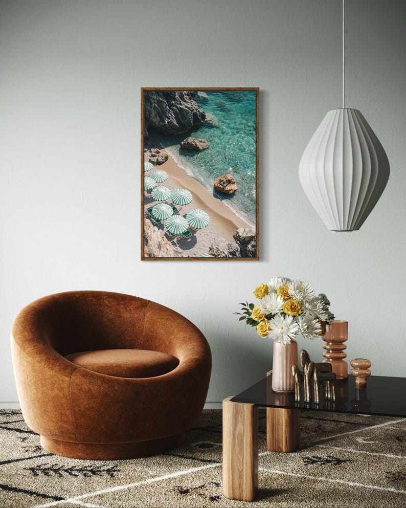 May include: A framed print of a beach scene with turquoise water, sandy shore, and green and white striped umbrellas. A brown velvet armchair, a black glass table, and a white pendant lamp are also visible in the room.