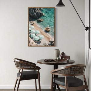 May include: A framed print of a beach scene with turquoise water, sand, and rocks. Several green and white striped umbrellas are on the beach. The print hangs above a small round table with two chairs. A black lamp is mounted on the wall.