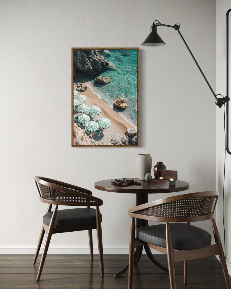 May include: A framed print of a beach scene with turquoise water, sand, and rocks. Several green and white striped umbrellas are on the beach. The print hangs above a small round table with two chairs. A black lamp is mounted on the wall.