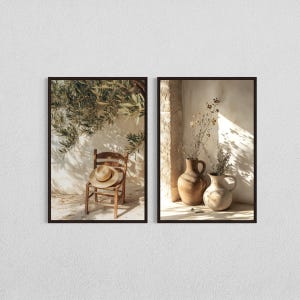 May include: Two framed prints. One features a wooden chair with a straw hat, set against a white wall with olive tree branches. The other shows two ceramic vases with dried plants, lit by sunlight.