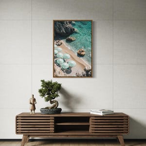 May include: A framed print of a beach scene with turquoise water, sand, and green and white striped umbrellas. The print is displayed above a wooden media console with a bonsai tree and a small statue.