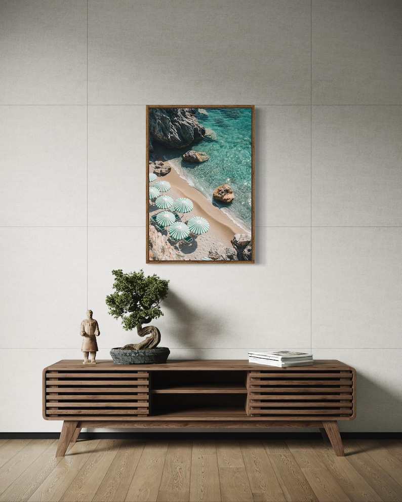 May include: A framed print of a beach scene with turquoise water, sand, and green and white striped umbrellas. The print is displayed above a wooden media console with a bonsai tree and a small statue.