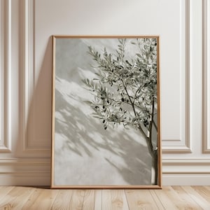 May include: A framed print depicting an olive tree branch with green leaves and black olives. The artwork is set against a light grey wall, with shadows adding depth. The frame is a natural wood colour, and the overall aesthetic is minimalist and natural.