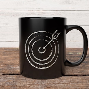 May include: Black coffee mug with a white target and arrow design. The target has concentric circles with an arrow in the center. The mug is on a wooden surface.