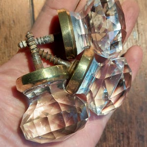 May include: Three clear glass cabinet knobs with brass bases. The knobs have a faceted design and are held in a hand.