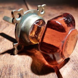 May include: A vintage amber glass door knob with a brass backplate and a decorative design. The knob is hexagonal in shape and has a slightly rounded edge.