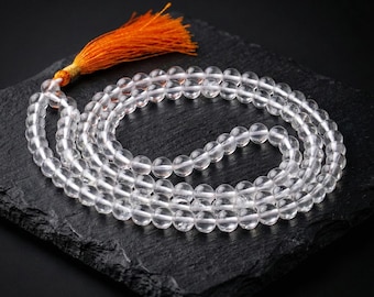 Op de afbeelding: Een ketting van heldere kwarts kristallen kralen met een oranje kwast. De ronde kralen zijn aan elkaar geregen, waardoor een gelaagd effect ontstaat. De ketting wordt gepresenteerd op een donker, getextureerd oppervlak.