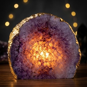 May include: A lit amethyst geode slice with a gold-plated edge. The interior glows with a warm, amber light, contrasting with the deep purple exterior. This decorative geode would be a unique addition to any home. The geode is a natural home decor item.