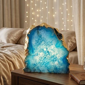 Pode incluir: Uma lâmpada de ágata azul e dourada em uma mesa de cabeceira de madeira. A lâmpada tem uma borda dourada e um interior azul. A lâmpada está acesa, com uma cama e luzes de corda ao fundo.