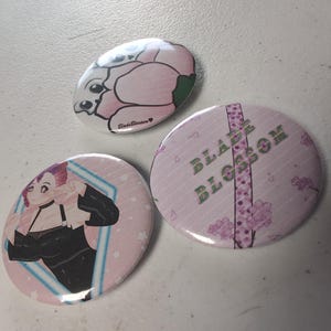 Puede incluir: Tres botones redondos. Uno presenta un personaje de dibujos animados rosa y blanco. Otro tiene un fondo rosa con una mujer con un atuendo negro. El tercer botón tiene el texto "BLADE BLOSSOM" con un diseño floral.
