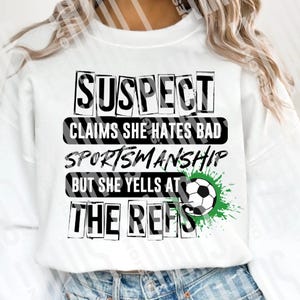 Pode incluir: Camisola branca com o texto "SUSPECT CLAIMS SHE HATES BAD SPORTSMANSHIP BUT SHE YELLS AT THE REFS" em letras pretas. Um gr&aacute;fico de bola de futebol com respingos de tinta verde est&aacute; &agrave; direita.