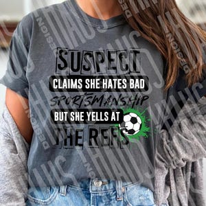 Pode incluir: Camiseta cinza com o texto preto "SUSPECT CLAIMS SHE HATES BAD SPORTSMANSHIP BUT SHE YELLS AT THE REFS". Um gr&aacute;fico de bola de futebol com detalhes verdes tamb&eacute;m est&aacute; presente na camiseta. A camiseta &eacute; usada com um cardigan cinza e jeans azuis.