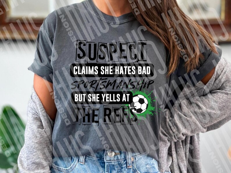 Pode incluir: Camiseta cinza com o texto preto "SUSPECT CLAIMS SHE HATES BAD SPORTSMANSHIP BUT SHE YELLS AT THE REFS". Um gr&aacute;fico de bola de futebol com detalhes verdes tamb&eacute;m est&aacute; presente na camiseta. A camiseta &eacute; usada com um cardigan cinza e jeans azuis.