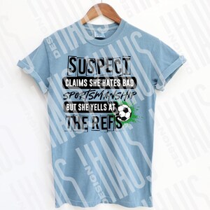 Pode incluir: Camiseta azul clara com o texto preto "SUSPECT CLAIMS SHE HATES BAD SPORTSMANSHIP BUT SHE YELLS AT THE REFS". Um gr&aacute;fico de bola de futebol com detalhes verdes est&aacute; no lado direito da camiseta. A camiseta est&aacute; pendurada em um cabide de madeira.