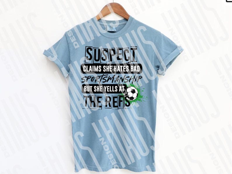 Pode incluir: Camiseta azul clara com o texto preto "SUSPECT CLAIMS SHE HATES BAD SPORTSMANSHIP BUT SHE YELLS AT THE REFS". Um gr&aacute;fico de bola de futebol com detalhes verdes est&aacute; no lado direito da camiseta. A camiseta est&aacute; pendurada em um cabide de madeira.