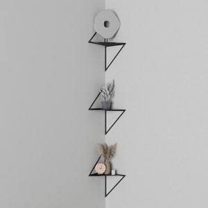 May include: Three black triangular shelves mounted in a corner of a white wall. The shelves are decorated with plants and a small clock.