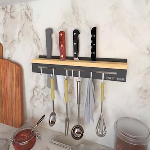 Op de afbeelding: Een keukenutensilienrek van zwart metaal en hout met haken om messen en keukengerei op te hangen. Het rek heeft een bovenste houten plank en een onderste zwarte metalen plank met haken. Het rek is gemonteerd aan een witte betegelde muur. De tekst "SWEET HOME" is op het rek gedrukt.