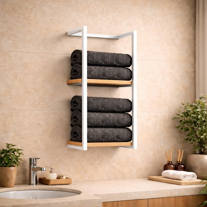 May include: A white metal and wood bathroom shelf unit with four rolled dark gray towels. The shelf has two wooden shelves and is mounted on a beige tiled wall. A sink and accessories are visible in the foreground.